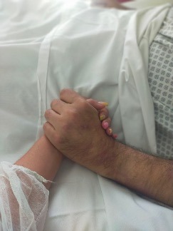 Two clasped hands on top of a person's torso, clothed in a hospital gown.