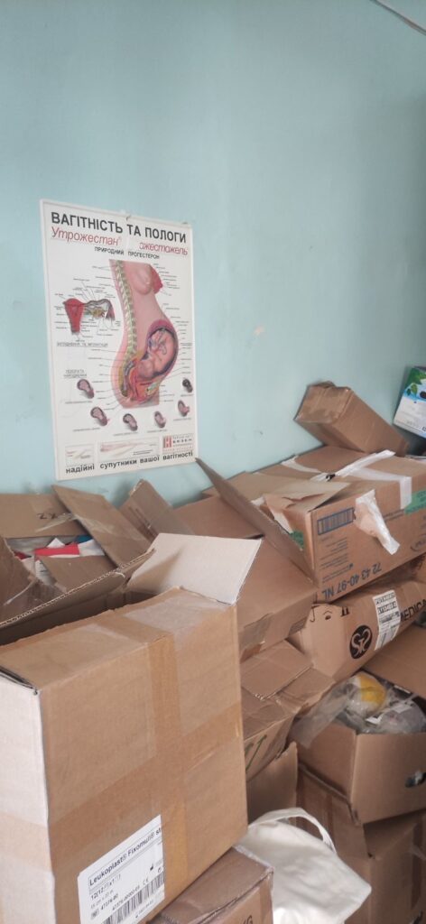 Cardboard boxes are stacked alongside a green wall, upon which hangs a medical poster depicting a pregnant woman's torso and uterus.