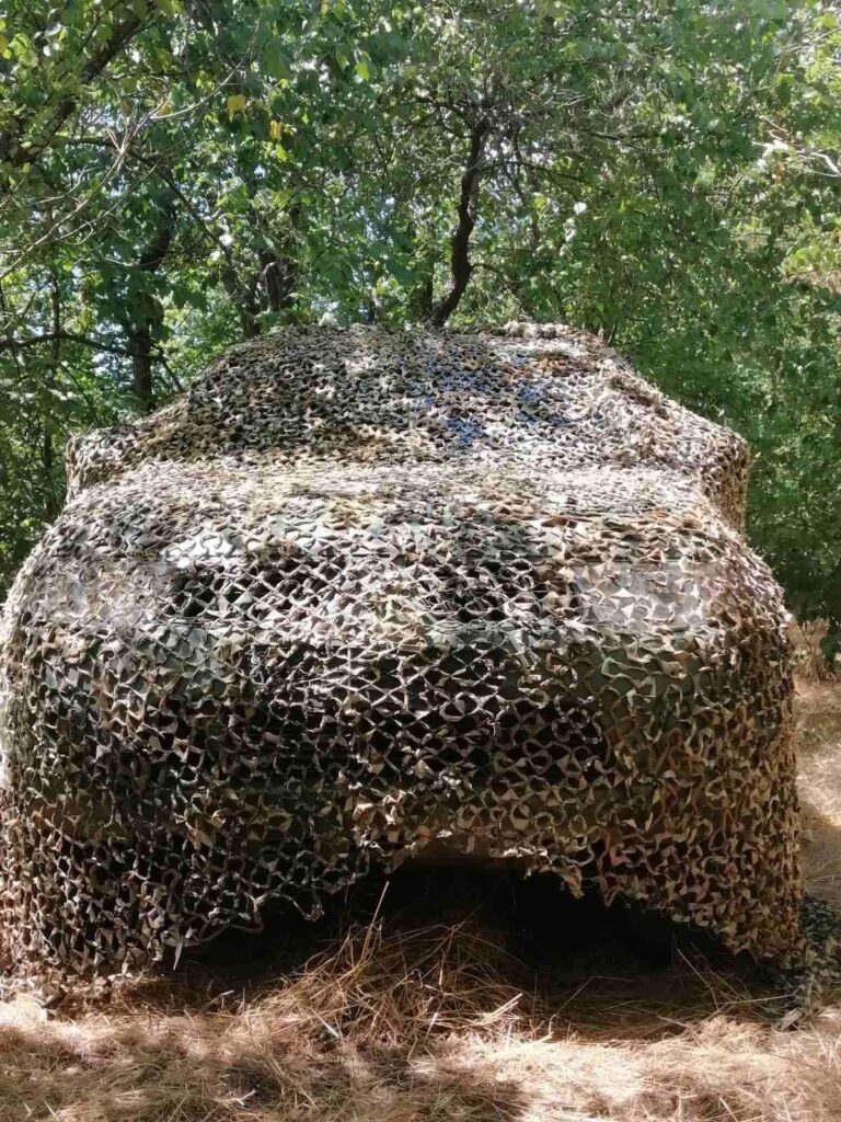 A camouflage net covers an army vehicle, next to green leafy trees.