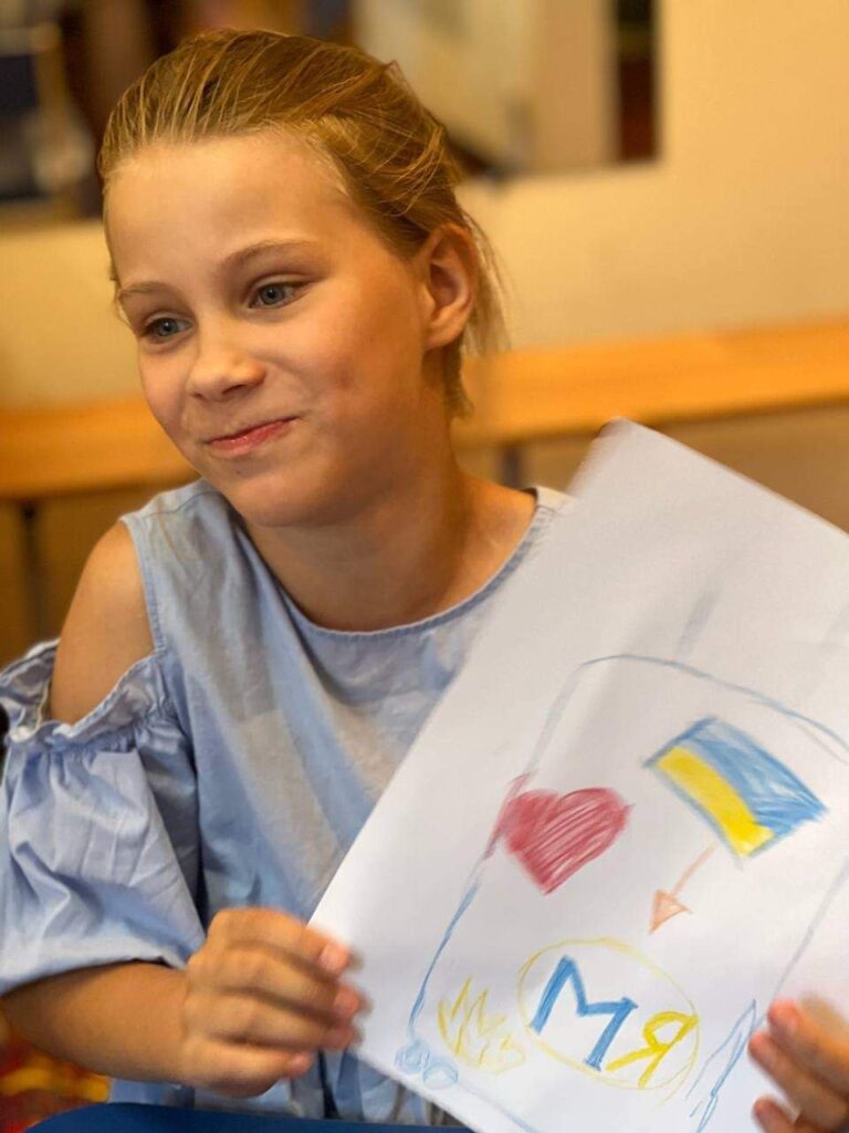 A girl, looking away from the camera, holds up a piece of paper with a drawing of a heart and a Ukrainian flag.