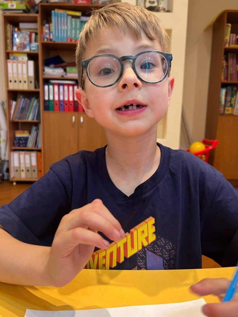 A boy wearing glasses sits at a desk with a piece of paper in front of him.