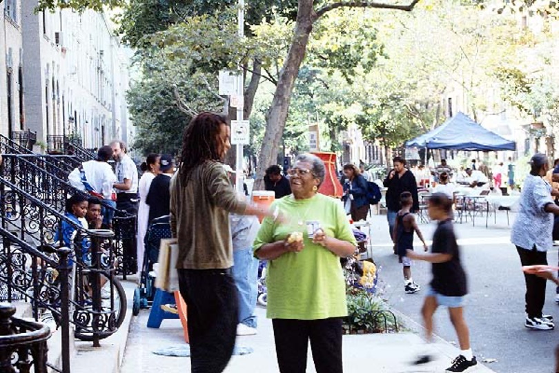 A picture of of a street in Harlem, New York.  A man and a woman are talking in the foreground, with several people in the background.  The street is closed off, and a tent and chairs can be seen.
