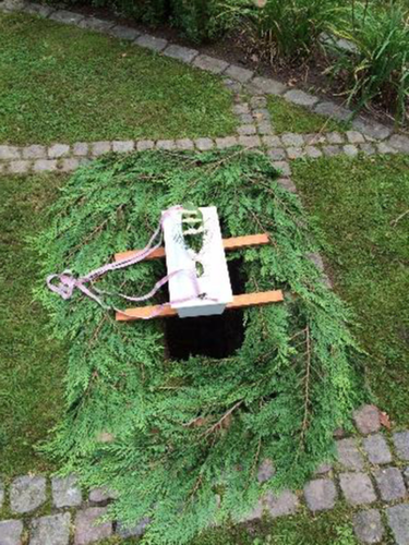 A small decorated white box suspended above a hole adorned with tree cuttings in a patch of grass.