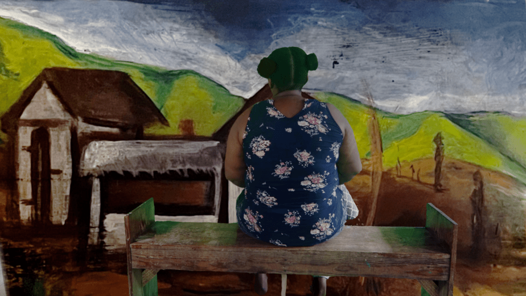 A woman sitting at a bench facing a mural of a colorful landscape with her back to the viewer.