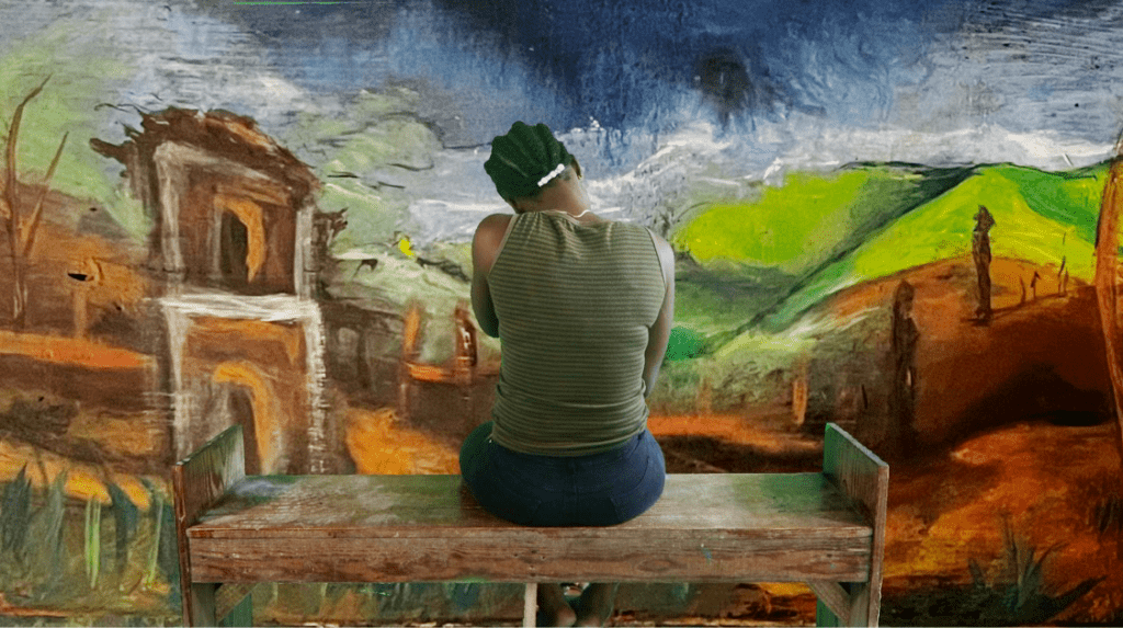 A woman sitting at a bench facing a mural of a colorful landscape with her back to the viewer.